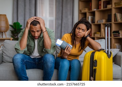 Cancelled Flight. Upset Black Couple After Tour Cancellation, Sitting On Sofa At Home With Travel Tickets And Suitcase. Vacation Problem And Restrictions For Tourists During Epidemic