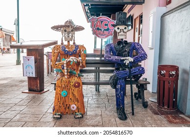 Cancun, Mexico. May 30, 2021. Skeleton Couple Sitting On Outdoor Bench At Roadside Against Entrance Of Restaurant