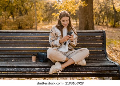 candid attractive young smiling woman sitting on bench in autumn park using phone wearing checkered coat, happy mood, fashion style trend - Shutterstock ID 2198106929