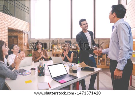 Candid of Indian creative design man smiling and shaking hands with coworker man or colleague and other clapping hand at office workplace. Introduction to team or marketing group concept soft tone.