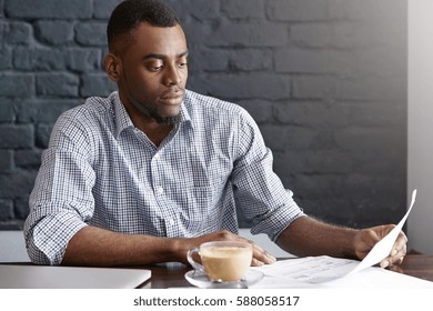 Candid Indoor Shot Of Attractive Dark-skinned Businessman In Checkered Shirt Holding Document And Reading Information With Concentrated Serious Look, Reviewing His Accounts During Lunch At Cafe