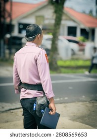 A candid, medium shot from a back view perspective capturing a male security officer or law enforcement personnel standing on duty by the roadside. The officer is wearing a light pinkish-purple uniform shirt with a red shoulder patch, black tactical pants, a duty belt, and a small utility pouch equipped with a flashlight on his hip. He is seen holding a dark blue digital tablet device in his hand, with a pair of smart glasses or a head-mounted device resting on the back of his head. The background is softly blurred (bokeh effect), showing a suburban street scene with tropical trees, vehicles, and a residential building with a red roof under broad daylight. This professional composition represents themes of public safety, law enforcement, security surveillance, community policing, modern security technology, and outdoor guard duty.