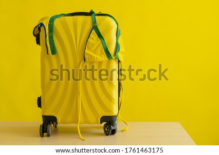 A candid mini-bikini of the coloring of the Brazilian flag hangs on a suitcase on a yellow background. The concept of a summer beach holiday. A woman is going on a trip to the sea.