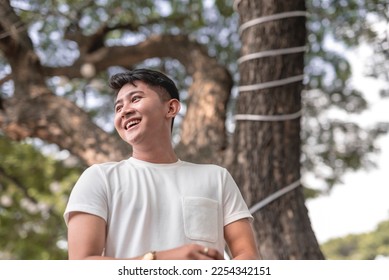 A candid photo of a happy and smiling young man wearing a plain white round neck shirt outside the park on a hot afternoon.