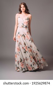 Candid portrait of a brunette young woman in summer gown with shoulder straps and floral print. Calm studio portrait of young lady in long sleeveless evening gown on grey background.