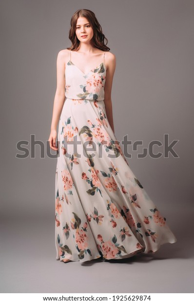Candid\
portrait of a brunette young woman in summer gown with shoulder\
straps and floral print. Calm studio portrait of young lady in long\
sleeveless evening gown on grey\
background.
