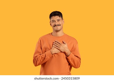 A candid portrait of a cheerful young man in an orange shirt, hands on heart, against a vibrant yellow background - Powered by Shutterstock