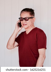 Candid Portrait Of Older Man In Red T Shirt And Sunglasses Using Smart Phone And Standing In Front Of White Wall