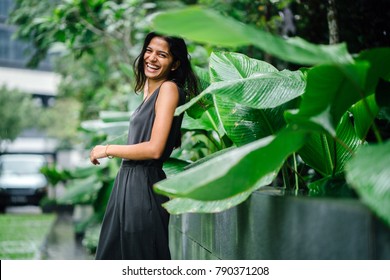 Candid Portrait Of A Young Indian Woman Standing In A Lush Green Garden And Laughing In A Natural, Authentic Way. She's Dressed In A Professional Grey Smock Jumpsuit. She Is Slim And Attractive.