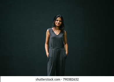 Candid Portrait Of A Young Woman Against A Plain Dark Background. She's Attractive And Dressed Professionally And Is Confident, Calm And Relaxed. Her Hands Are In Her Pockets. 