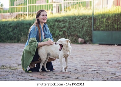 Candid Portrait Of Young Woman Kneeling Outside On Stone Ground, Covered With Blanket, Holding Stray Dog Yawning Next To Her