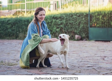 Candid Portrait Of Young Woman Kneeling Outside On Stone Ground, Covered With Blanket, Caressing Stray Dog Yawning Next To Her