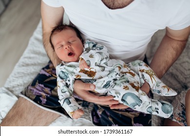 Candid Real Family Moment. Soft Focus. Young Dark Hair Father Holding His Newborn Baby Son Sitting In A Bedroom. Boy Is Wearing Body With Watercolor Animal Print. Father's Day. Boy Is Yawning.