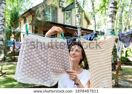 Candid real life portrait of young adult beautiful attractive caucasian woman hanging up fresh washed family clothes on birch tree clothesline with pins at home yard on bright sunny day outdoors