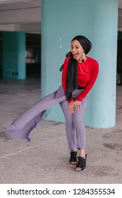 A Candid And Relaxed Moment Of A Muslim Malay Woman Laughing Out Loud. She Is Bending Over Slightly As She Stands. The Woman Is Wearing A Fashionable Minimalist Outfit And A Tudung Head Scarf Hijab. 