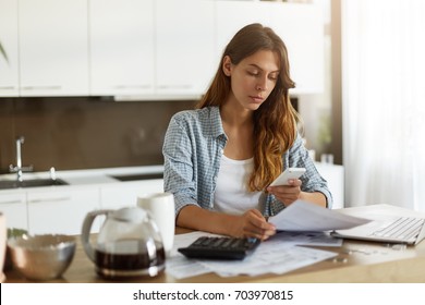 Candid shot of beautiful concentrated young Caucasian woman typing text message on mobile phone while calculating bills in kitchen, sitting at table with laptop computer, papers and calculator - Shutterstock ID 703970815