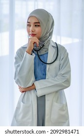 Candid Shot Of Beautiful Muslim Islam Arabian Female Professional Intern Doctor Practitioner In White Lab Coat Hijab And Stethoscope Standing Smiling Thinking Holding Hand At Chin Crossed Arm Alone.