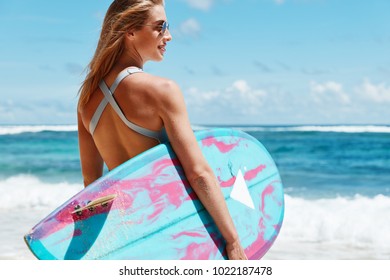 Candid shot of blonde female in bathing suit, has tanned healthy skin, holds bodyboard under arm, waits for friend, going to surf and hit waves together, spends unforgettable vacations on island - Powered by Shutterstock