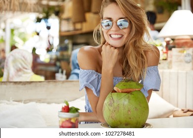 Candid Shot Of Good Looking Fashionable Woman In Sunglasses, Has Broad Smile With White Perfect Teeth, Sits Against Cafe Background, Drinks Coconut Cocktail, Enjoys Unforgettable Summer Holidays