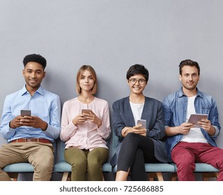 Candid Shot Of Group People Meet Accidentally In One Office, Sit At Comfortable Chairs, Use Cell Phones And Table Computer, Communicate. Office Workers In Coworking Space, Isolated Over Grey Wall