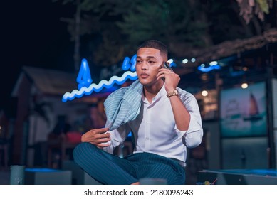 Candid Shot Of A Handsome And Dapper Young Man Hanging Out At An Outdoor Bar. Holding A Phone Listening To Someone. Nightlife Scene At A Popular Spot.