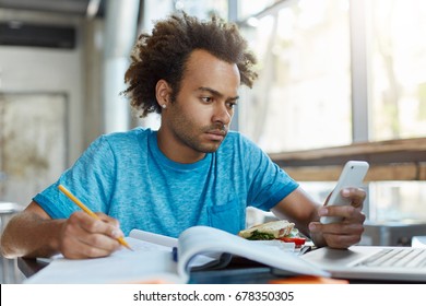 Candid Shot Of Handsome Stylish Black Student Of Law School Writing Down In Copybook While Doing Home Assignment At Empty Coffee Shop, Holding Cell Phone And Reading Something With Serious Look