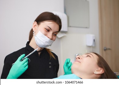 Candid Shot Of Positive Professional Female Dentist Wearing Mask And Gloves Holding Syringe, Going To Anesthetize Tooth Nerve Of Smiling Woman Patient Before Dental Treatment. Anesthesia And Numbing