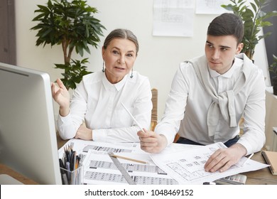 Candid Shot Of Stylish Experienced Gray Haired Senior Female Architect Pointing At Computer Screen, Explaining Point Of View To Her Young Male Colleague While Working On New Housing Project Together,