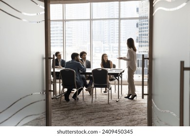 Candid shot through doorway of diverse business team and female leader discussing teamwork at large table. Group of young entrepreneurs negotiating, networking in modern office meeting room - Powered by Shutterstock