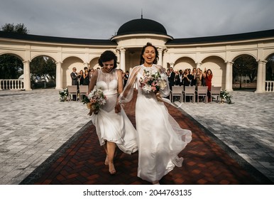 Candid Shot Of Two Female Lesbian LGBT Brides Walking Away From The Wedding Officiant, Guests Cheering