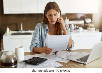 Candid shot of worried young European female dressed casually sitting at kitchen table, holding paper sheet and talking to house and utilities service representative about miscalculation in bills