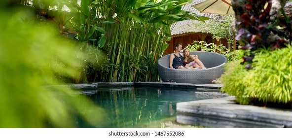 Candid Shot Of Young Caucasian Couple Lounging In Modern Chair Near Luxurious Pool Of Tropical Hotel And Spa In Bali