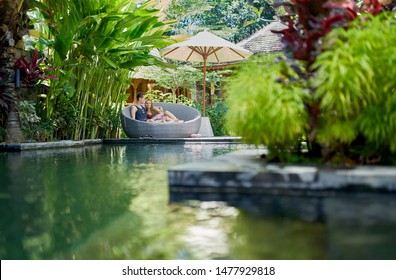 Candid shot of young caucasian couple lounging in modern chair near luxurious pool of tropical hotel and spa in Bali - Powered by Shutterstock