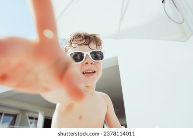 A candid shot of a young child joyfully reaching towards the camera, sporting white sunglasses, capturing the essence of a sunny summer vacation day. - Powered by Shutterstock
