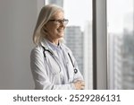 Candid smiling mature female doctor modern private medical center leader stand by window look at distance make ambitious plans visualize successful work feel happy for perfect care of patients health