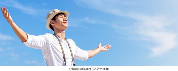 Candid Of Young Attractive Asian Man Close Eyes, Deep Breath And Wide Open Arms In Blue Sky View Background Feeling Wind And Sunshine. Happy Asian Hipster Male In Youth Freedom Culture Travel Concept.