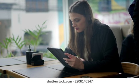 Candid Young Woman Sitting At Coffee Shop Reading Content Online On Tablet Device. Person Paying Attention To Screen Using Modern Technology