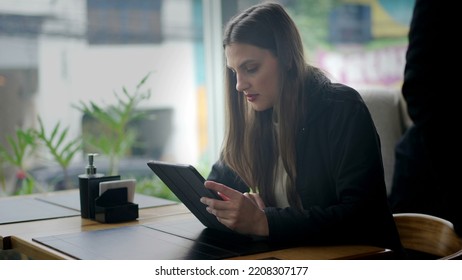 Candid Young Woman Sitting At Coffee Shop Reading Content Online On Tablet Device. Person Paying Attention To Screen Using Modern Technology