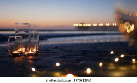 Candle flame lights in glass, romantic beach date, California ocean waves, sea water. Candlelight seamless looped cinemagraph. Wineglass on sand, garland in twilight dusk. Illuminated pier reflection.
