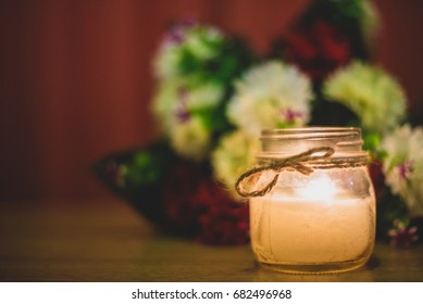 Candle In A Jar In Front Of Flower Bouquet, Decorative For Bridal Themes Or Sad Occasions