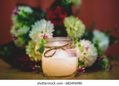 Candle In A Jar In Front Of Flower Bouquet, Decorative For Bridal Themes Or Sad Occasions