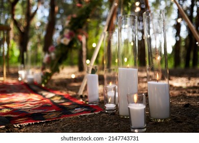Candles burning in glass vases, flasks on carpet near bohemian tipi arch decorated with roses and flowers wrapped in fairy lights on outdoor wedding ceremony venue in pine forest. Boho rustic decor.