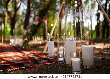 Candles burning in glass vases, flasks on carpet near bohemian tipi arch decorated with roses and flowers wrapped in fairy lights on outdoor wedding ceremony venue in pine forest. Boho rustic decor.