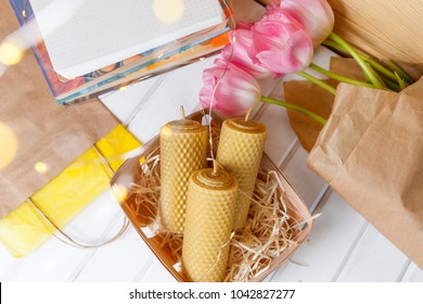 Candles From The Honeycomb Lie In A Basket Near Pink Lulips, Books, Packet And Fairylights On The Background Of A Wooden Table With Yellow Boke Shot From Above.