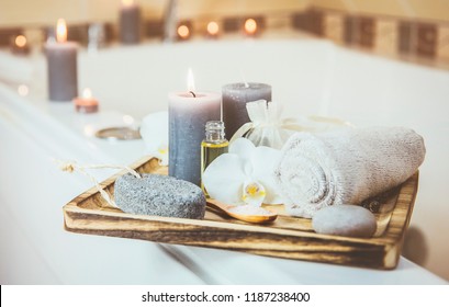 Candles and spa essentials on wooden tray in bathroom, essential oil, bath salt on wooden spoon, pumice stone, towel, sea stone, white orchids. Spa products set conception. Instagram filter style.