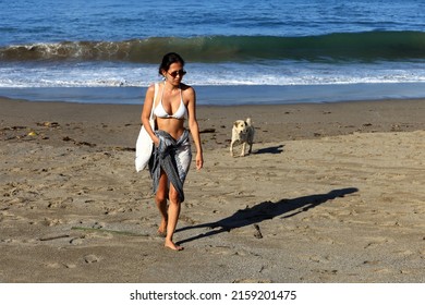 Canggu, Bali, Indonesia - March 17, 2022.
A Young Asian Woman Wearing A White Bikini And A Sarong Walking Up The Beach With A Dog Behind Her At Batu Bolong, Canggu, Bali.