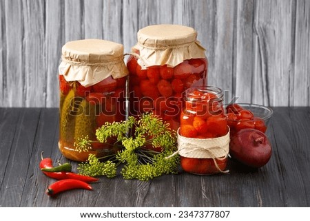 Canned cherry tomatoes and cucumbers in jars and a bowl, hot chili peppers, onions on a wooden background. Homemade preserves, pickled vegetables.
