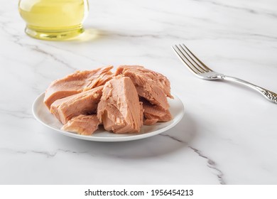 Canned Tuna Fillet Pieces On A White Saucer And Fork Over Marble Table Close-up. Preserved Tuna Fish Meat As Low Calorie Ingredient For Dieting And Healthy Eating. Tasty Seafood. Front View.