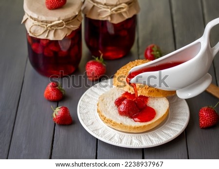 Canning Strawberries. Adding homemade strawberry syrup on sliced white bread bun with portion of whole strawberry berries on it. Selective focus. Glass jars with canned strawberries on background.