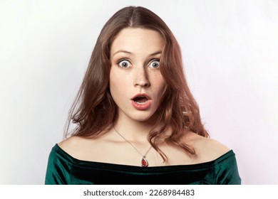 I can't believe this. Portrait of shocked astonished woman wearing green dress, looking at camera with big eyes and open mouth, being very surprised. Indoor studio shot isolated on gray background.